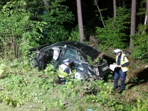Zdjęcie policjantów sporządzających dokumentację na miejscu wypadku w tle widać las i rozbity samochód.