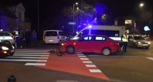 Zdjęcie przedstawiające skrzyżowanie z przejściem dla pieszych i przejazdem rowerowym na którym leży rower obok dwaj policjanci oraz dwa radiowozy i czerwone auto które potrąciło rowerzystę.