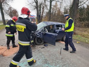Strażacy i policjant przy uszkodzonym aucie w tle karetka pogotowia.