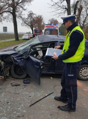 Policjant obok samochodu biorącego udział w wypadku trzymający w ręku notatnik służbowy.