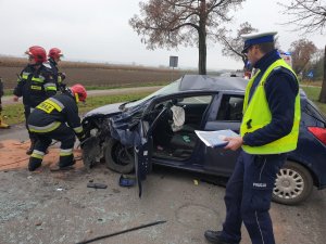 Strażacy podczepiający linkę holowniczą do samochodu obok policjant.