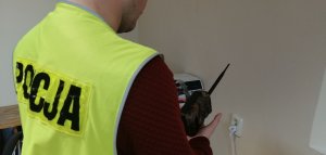 zdjęcie przedstawiające policjanta trzymającego w ręku aparat fotograficzny, który mocuje się do drzewa w celu robienia zdjęć w lesie głównie migrującym zwierzętom tak zwaną fotopułapkę.