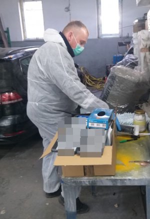 zdjęcie przedstawia technika kryminalistyki ubranego w biały kombinezon i maseczkę który zbiera ślady z tablic rejestracyjnych w garażu w którym znajduje się odzyskane z kradzieży auto na dalszym planie samochód koloru czarnego którym poruszali się sprawcy kradzieży