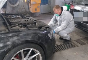 zdjęcie przedstawia technika kryminalistyki ubranego w biały kombinezon i maseczkę klęcząc zbiera ślady z tablic rejestracyjnych przymocowanych do odzyskanego z kradzieży samochodu. Czynności prowadzone są w garażu w którym znajduje się odzyskane z kradzieży auto na dalszym planie samochód koloru czarnego którym poruszali się sprawcy kradzieży