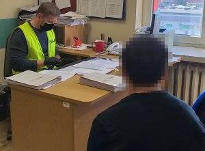 zdjęcie z przesłuchania jednego ze sprawców kradzieży samochodu policjant siedzi przed biurkiem i notuje w komputerze zeznania zatrzymanego który siedzi tyłem do zdjęcia . Na biurku widać rozłożone teczki z dokumentami.
