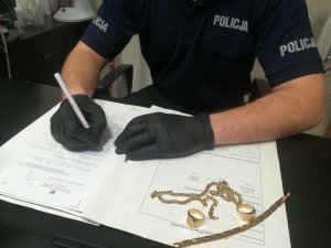 na zdjęciu widać dłonie funkcjonariusza siedzącego przy biurku który sporządza dokumentację  obok dokumentów leżą rzeczy z przestępstwa czyli fałszywe złoto dwa sygnety, bransoletka i łańcuszek