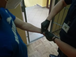 na zdjęciu widać jak policjant, który ma na dłoniach rękawiczki zdejmuje kajdanki osobie ubranej w niebieską koszulkę