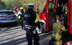 na zdjęciu stoją dwaj policjanci po lewej policyjny radiowóz po prawej wóz strażacki na stopniu którego siedzi sprawca wypadku opatrywany przez kucającego przy nim strażaka, w tle widać las.