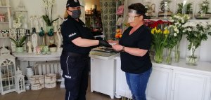 zdjęcie zrobione w kwiaciarni na półkach i kwietnikach jest bardzo dużo różnego rodzaju kwiatów i ozdób do domu, policjantka wręcza właścicielce ulotki dotyczące właściwego zachowania się gdy podejrzewamy zarażenie się koronawirusem obie mają na sobie maseczki ochronne