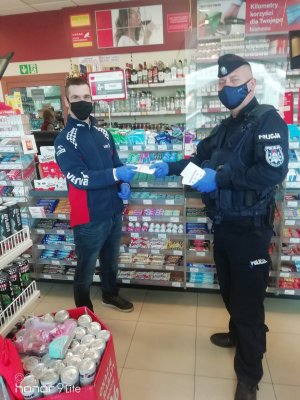 umundurowany policjant wręcza pracownikowi stacji paliw ulotki , obaj mają ubrane maseczki i rękawiczki, w tle widać półki z towarem
