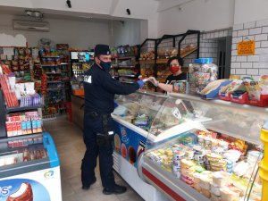umundurowany policjant który ma na twarzy maseczkę ochronną a na rekach rękawiczki przekazuje ulotki ekspedientce w sklepie spożywczym.