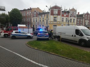 zdjęcie z miejsca zdarzenia po prawej samochód dostawczy po lewej wóz strażacki a po środku dwa radiowozy policyjne obok nich policjanci i strażacy w dalszym planie widać kamienicę i drugą karetkę.