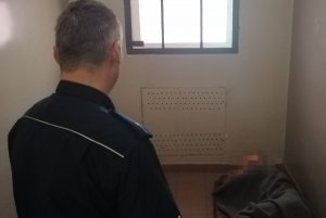 Zdjęcie przedstawia policjanta stojącego przy otwartych drzwiach celi a na materacu lezący mężczyzna zakryty kocem  który był zatrzymany do sprawy włamań