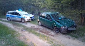 na zdjęciu leśna droga na której stoją dwa samochody. Na pierwszym planie stoi fiat koloru zielonego z rozbitym przodem, a tuż za nim radiowóz policyjny . Zielonym fiatem uciekał przed kontrolą policyjną osiemnastolatek który jechał nim bez uprawnień.