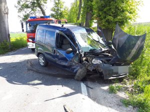 na tym zdjęciu widać przód niebieskiego samochodu renault stojącego przodem na poboczu , widoczne są na nim uszkodzenia wynikające z uderzenia w drzewo . Za uszkodzonym samochodem widać czerwony wóz strażacki. MIejsce wypadku otaczają drzewa posadzone wzdłuż ulicy.