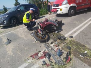 zdjęcie policjanta kucającego przy rozbitym na środku ulicy motocyklu, na około widać jego połamane elementy rozlane płyny techniczne po prawej stronie stoi wóz strażacki a na wprost samochód w ciemnym kolorze który uderzył w motocyklistę