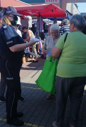 policjantka wręczająca na targowisku miejskim innej kobiecie maseczkę ochronną w celu zakrywania ust i nosa w tle widać stragany i ludzi przemieszczających się po targowisku