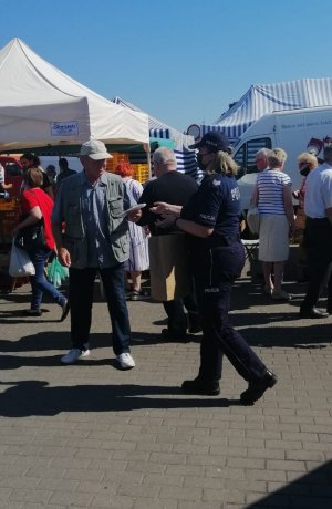 policjantka wręczająca na targowisku miejskim mężczyźnie maseczkę w tle widać stragany i spacerujących ludzi