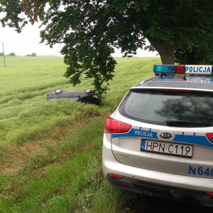 zdjęcie radiowozu stojącego na poboczu przed nim drzewo a po lewej stronie w polu z rzepakiem samochód na dachu