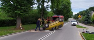 ulica na której widać przewrócone na dach auto w kolorze żółtym obok niego przechodzi policjant , z przewróconym samochodem stoi wóz strażacki i grupa strażaków po prawej stronie zaparkowany biały samochód