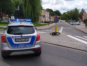 zdjęcie wykonane na skrzyżowaniu na pierwszym planie stoi radiowóz z włączoną sygnalizacją błyskową na wysepce która jest na skrzyżowaniu widać przewrócony rower w tle widać budynki