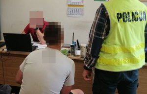 na zdjęciu widać siedzącego na krześle zatrzymanego mężczyznę obok niego stoi funkcjonariusz który ma założoną kamizelkę odblaskową naprzeciw przy biurku siedzi inny policjant którzy przesłuchuje sprawcę kradzieży łańćuszka