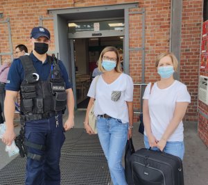 zdjęcie zrobione przed wejściem do sklepu widać na nim policjanta obok dwóch kobiet , wszyscy mają założone maseczki policjant w ręku trzyma maseczki w celu rozdania klientom sklepu, jedna z kobiet ma w ręku walizkę