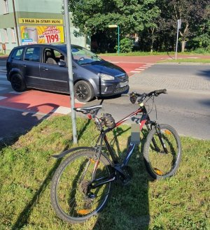 zdjęcie przedstawia fragment jezdni i przejścia dla pieszych na którym stoi zaparkowany samochód koloru czarnego który potrącił rowerzystę, rower stoi opary o znak drogowy na poboczu w tle widać budynki