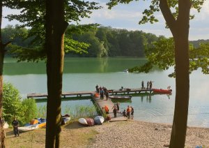na zdjęciu widać jezioro i pomost jest na nim bardzo dużo ludzi między innymi strażaków i policjantów a także osoby postronne, na jeziorze pływa łódka straży pożarnej na brzegu widać łódki i kajaki