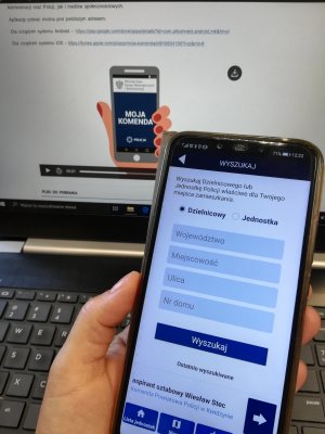 zdjęcie dłoni w której trzymany jest telefon z włączoną aplikacją moja komenda w tle widać włączony laptop również z aplikacją moja komenda