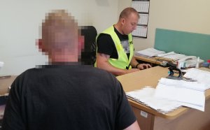 zdjęcie przedstawia zatrzymanego mężczyznę siedzącego tyłem do fotografa przed nim siedzi przy biurku przesłuchujący go policjant który ma na sobie żółta kamizelką z napisem policja, na biurku są porozkładane materiały do prowadzonych spraw