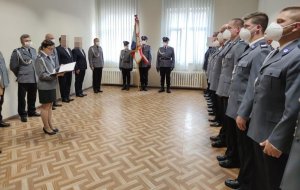 zdjęcia przedstawia ustawionych po prawej stronie w szeregu policjantów naprzeciw nich stoi kobieta która odczytuje pismo za nią stoją inni funkcjonariusze  na wprost od fotografa stoi poczet sztandarowy składający się z dwóch mężczyzn i kobiety środkowy policjant trzyma w ręku sztandar