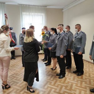 zdjęcia przedstawia funkcjonariuszy stojących po prawej stronie w dwuszeregu kilku mężczyzn i kobietę wszyscy są ubrani w mundur odświętny tak zwaną gabardynę. Komendant policji wręcza policjantce słonecznik i akt mianowania obok niego stoją dwie kobiety , w tle widać poczet sztandarowy