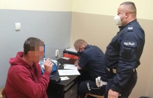 na zdjęciu widać dwóch policjantów, jeden siedzi i sporządza dokumentację drugi stoi i przeprowadza badanie alkomatem osoby zatrzymanej która jechała samochodem będąc pod działaniem alkoholu, zatrzymany mężczyzna ubrany w czerwoną bluzę z kapturem siedzi na krześle ma w ręku rurkę z ustnikiem w którą wdmuchuje powietrze.