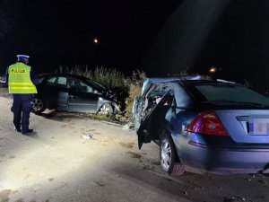 zdjęcie zrobione po zmroku jest ciemno, na poboczu ulicy widać dwa samochody rozbite a pośrodku idzie policjant który będzie robił oględziny obu aut.