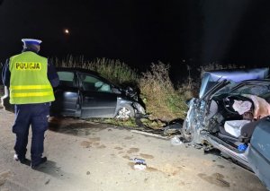 podobne zdjęcie do poprzedniego, zmrok na poboczu ulicy dwa rozbite auta a pomiędzy nimi policjant sporządzający dokumentację z miejsca zdarzenia.