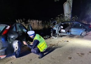 na dworze zmrok na poboczu ulicy dwa rozbite auta za nimi drzewo a pomiędzy nimi policjant który klęczy i opisuje uszkodzenia auta , policjant sporządza dokumentację z miejsca zdarzenia.