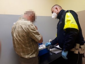 zdjęcia przedstawia policjanta w pomieszczeniu przejściowym, funkcjonariusz ubrany jest w niebieski mundur ma na ramionach założone odblaski , jest to policjant wydziału ruchu drogowego który bada alkomatem mężczyznę zatrzymanego za jazdę pod wpływem alkoholu.