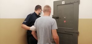zdjęcie przedstawia umundurowanego funkcjonariusza który wprowadza do pomieszczenia dla osób zatrzymanych mężczynę ubranego w siwą koszulkę