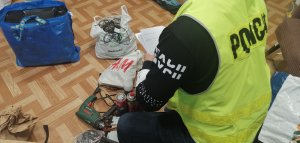 na zdjęciu widać policjanta ubranego w cywilne ubranie który ma założoną kamizelką odblaskową z napisem policja . Policjant kucając przy odzyskanych z kradzieży m.in.elektronarzędzia spisuje protokół
