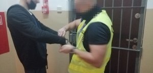na zdjęciu widać dwóch mężczyzn z zamazanymi twarzami gdzie jeden jest policjantem ubranym w żółtą kamizelkę odblaskową z napisem policja a drugi to zatrzymany mężczyzna któremu funkcjonariusz zakłada kajdanki. W tle widać ścianę i kratę przy drzwiach wejściowych do pomieszczenia dla osób zatrzmanych