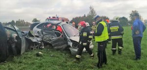 zdjęcie z wypadku drogowego, na pierwszym planie mężczyzna, policjant ruchu drogowego spisujący dane uczestników wypadku w dalszej części zdjęcia widać uszkodzone samochody. Obok nich widać straż pożarną oraz dwóch ratowników medycznych gdzie jeden jest w uszkodzonym aucie i udziela poszkodowanej kobiecie pomocy. W tle widać wóz strażacki oraz osoby postronne.