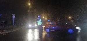 zdjęcie zrobione wieczorową porą widać na nim skrzyżowanie na którym stoi policjantka kierująca ruchem , obok zgodnie z poleceniem widać dwa auta które jadą we wskazanym przez nią kierunku.