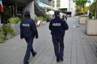 zdjęcie przedstawia dwie funkcjonariuszki idące chodnikiem po lewej stronie widać duży budynek i schody na wprost od policjantek spacerują ludzie po  prawej stronie betonowe kwietniki z  krzewami.