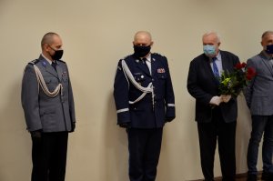 zdjęcie przedstawia czterech mężczyzn stojących przy ścianie obok siebie dwóch z lewej jest w mundurze a dwóch następnych w garniturze, wszyscy mają założone maseczki na twarz . Jeden z mężczyzn stojący w garniturze trzyma w dłoni kwiaty, róże.