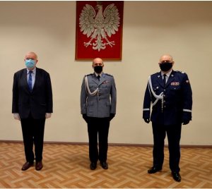 zdjęcie zrobione w świetlicy Komendy Powiatowej Policji w Kwidzynie w tle jasna ściana na której wisi Godło Polski czyli biały orzeł na czerwonym tle, na pierwszym planie stoi trzech mężczyzn jeden w garniturze a dwóch w mundurze policyjnym.