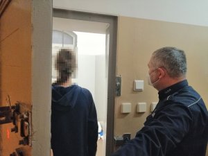 zdjęcie przedstawia policjanta stojącego przed celą dla osób osadzonych. W celi widać  kobietę, która jest zatrzymana do sprawy dotyczącej posiadania narkotyków.