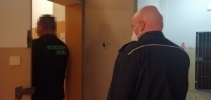 zdjęcie przedstawia policjanta otwierającego drzwi do celi w progu której stoi mężczyzna, zatrzymany do sprawy znieważenia i naruszenia nietykalności cielesnej funkcjonariusza policji podczas przeprowadzania interwencji oraz jazdy samochodem pod wpływem alkoholu.