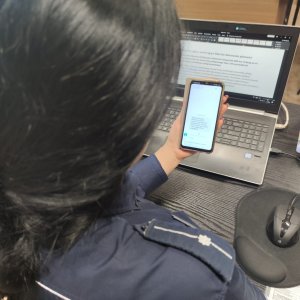 zdjęcie siedzącej przed komputerem policjantki trzymającej w ręku telefon z fałszywym sms-em w którym jest informacja o tym aby dokonać płatności za niewymiarową przesyłkę kurierską