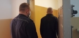 zdjęcie przedstawia funkcjonariusza stojącego przy otwartych drzwiach do celi w pomieszczeniu dla osób zatrzymanych, przed nim stoi mężczyzna w czarnej bluzie, który jest zatrzymany do sprawy narkotykowej